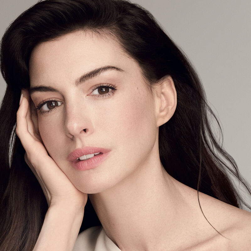 The actress Anne Hathaway holding a closed jar of Shiseido Vital Perfection Uplifting and Firming Advanced Cream against a white background. The closed jar features the Shiseido logo, product name, and the words "ReNeuraRED Technology." 