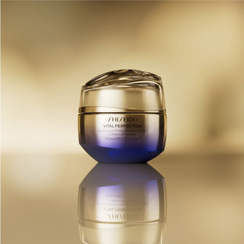 A  round, closed gold-and-purple-colored jar of Shiseido Vital Perfection Uplifting and Firming Advanced Cream. The jar is shown against a blurred gold background.  